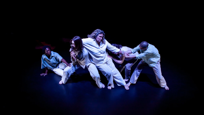Four dancers perform in darkness, all crouching down. The dancer in the middle leans on the others. One dancer is sat, watching from behind them. 