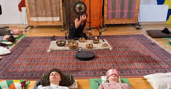 Neeta hosting a sound bath ar open source arts. She is ringing tibetan singing bowls sat on a rug, attendents are all lying down, eyes closed listening