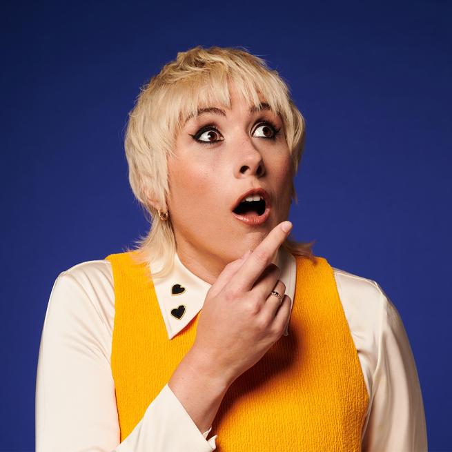 With a shocked expression and bleached blonde hair in a wide-collared shirt, Maisie Adam looks wide-eyed and open-mouthed as she points off-camera.
