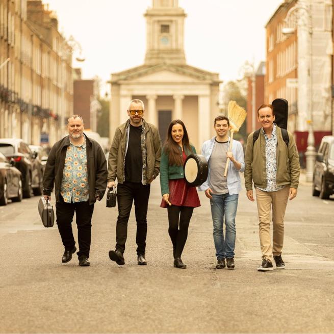 On a sunny street, the members of Irish House Party smile and walk towards the camera, each carrying an instrument case, one holding a broom.