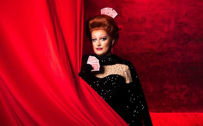 Kate Butch stands behind a red curtain, she holds a pack of cards.