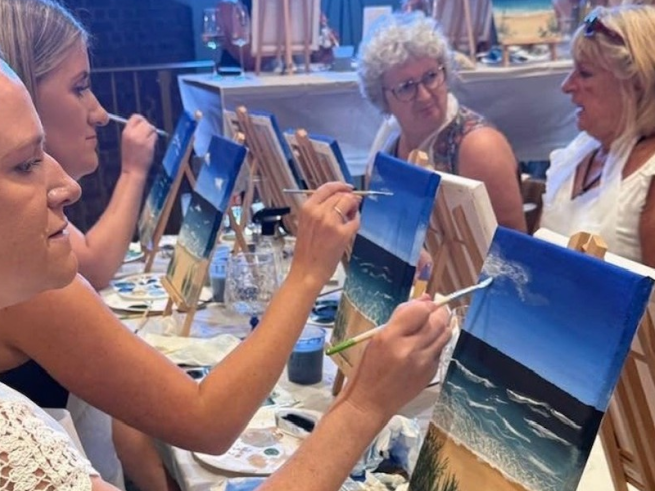 People of various ages taking part in a relaxed Sip & Paint session, painting blue coastal scenes on canvases at easels while chatting together around a table.