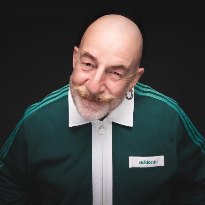 Jeff Innocent, a bald man with a beard and moustache, leans towards the camera, smirking. He wears a dark green Adidas tracksuit with a white collar.