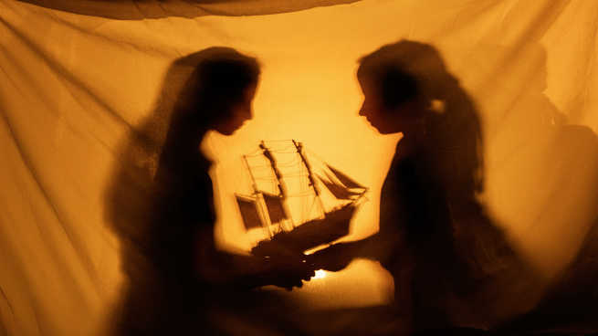 The outline of two young people in a tent or behind a curtain, holding a miniature ship.