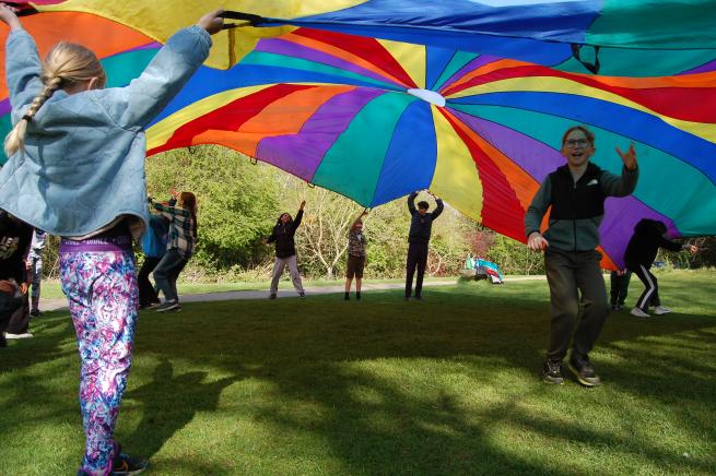 A group of children playing a parachute game with a rainbow striped parachute.