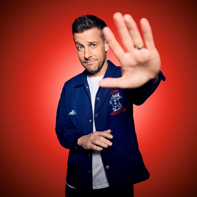 Chris Ramsey with his palm up to the camera smirking. 