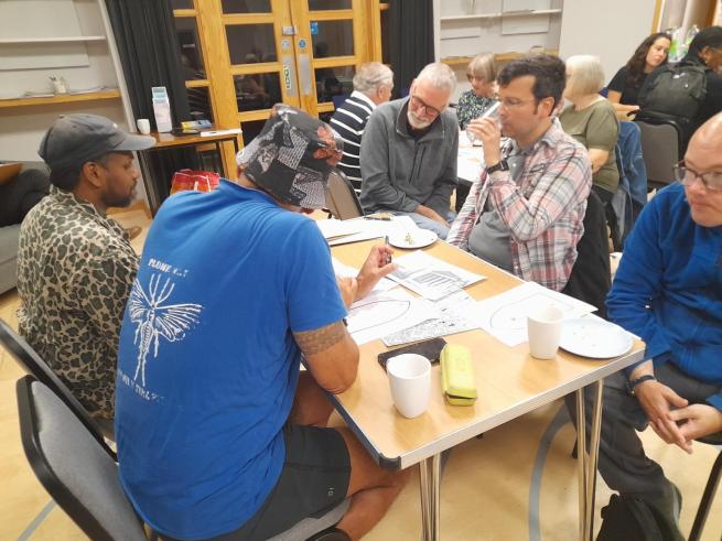 A group of people sit at a table, taking part in discussion. The table has papers, mugs and plates on it from lunch.