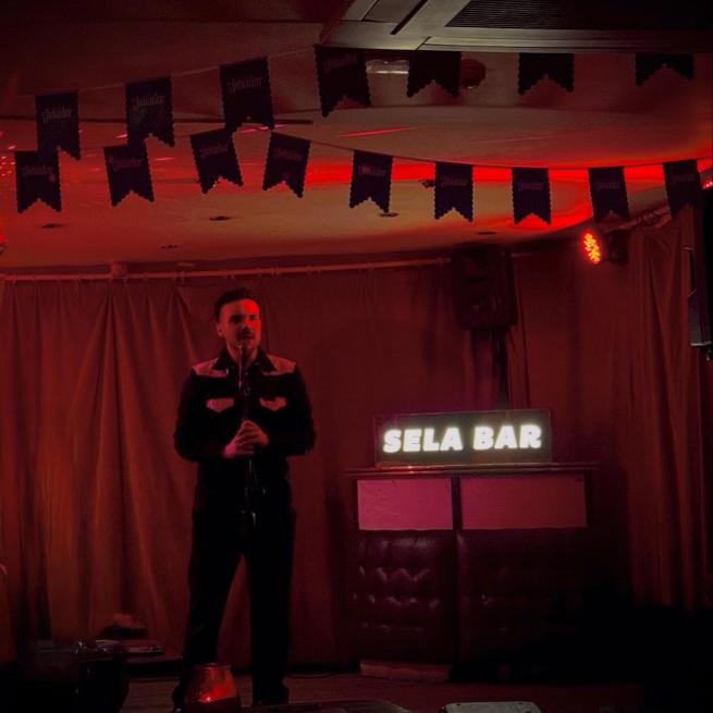 A white man with dark hair, a moustache and a jazzy black and white shirt talks into a microphone on a red lit stage. 