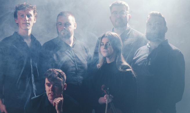 Six musicians stand, except for one that seems to be sat or squatting, it's hard to tell with all the smoke, look like  good band though