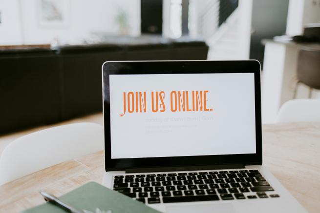 Image shows a laptop on a desk, next to a pad of paper and pen. The laptop screen reads "Join us online". In the background, there is a sofa and open plan rooms.