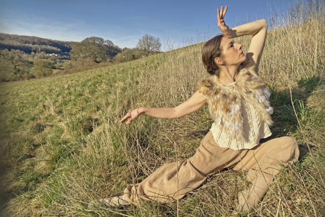 A dancer in beige poses on a grassy hillside