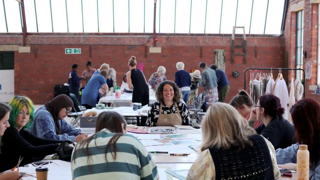 A workshop teacher smiling into the camera from a large workshop table filled with participants, paper and craft materials. The workshop is taking place on the top floor of the 1912 Mill - a red brick heritage Mill building with high ceilings and skylights.