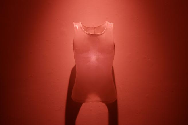 A translucent sleeveless vest hangs against a deep red wall illuminated by red light. 