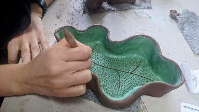 A hand is holding an instrument to carve into clay. The sculpture appears to be a deep dish in the shape of an oak leaf.