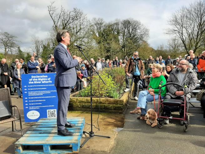A man standing on a raised platform addressing a crowd. There is a notice behind him saying 'Declaration of the Rights of the Rover Wharfe'