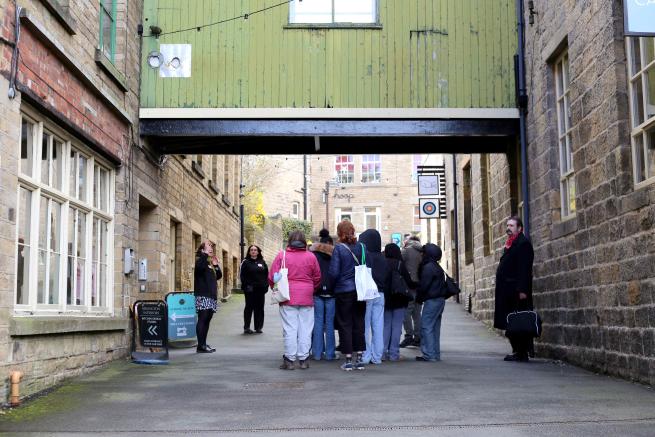 A group of visitors on a Heritage Tour of Sunny Bank Mills. They are gathered on the Mills' main hill under a green painted wooden clad bridge, connecting two Yorkshire stone built mill buildings.