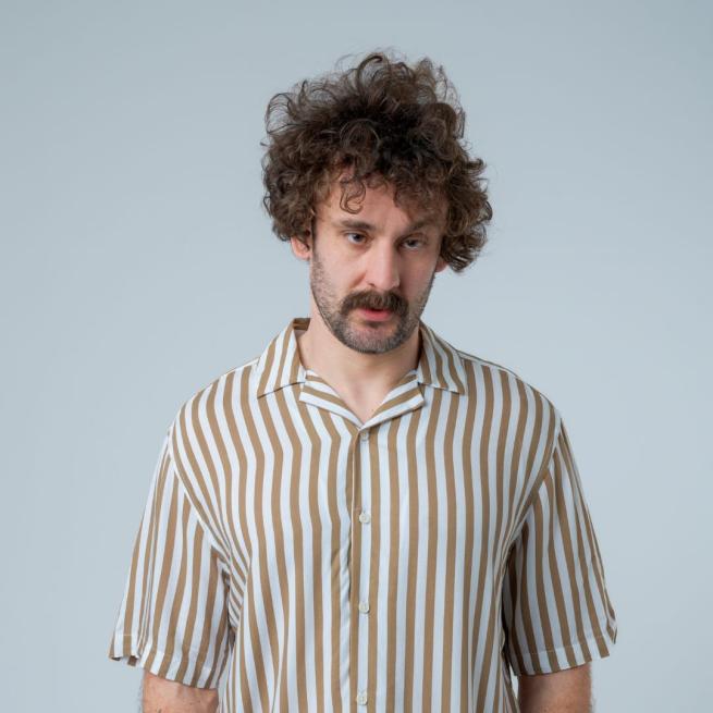 Josh Pugh stands with a neutral gaze and characteristic messy hair, wearing a striped shirt.