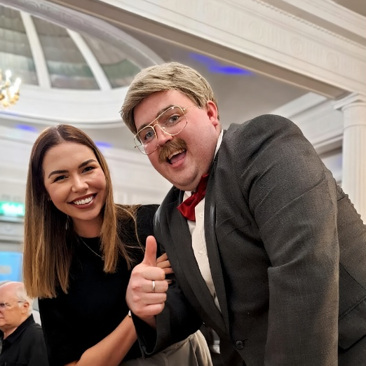 A photograph of a Brian Potter from Phoenix Nights impersonator having a photograph with a young female fan during a comedy dinner show. They are smiling and he has his thumb up.