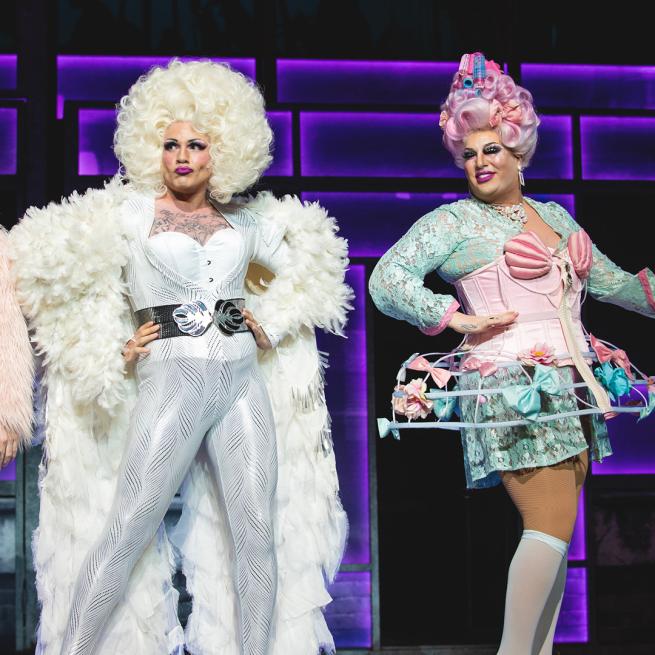 Two drag performers in elaborate outfits and bouncy wigs pose candidly on stage.