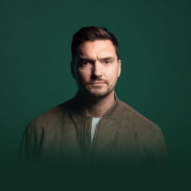 Against a dark green backdrop, Jack Skipper stands in a brown jacket and holds a neutral expression to the camera.