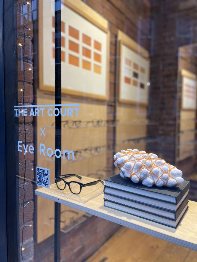 Looking into a shop window there is a vinyl sign saying THE ART COURT X Eye Room with a QR code beneath in. Inside we can see a light wooden shelf with a pair of thick rimmed glasses and a stack of books with a lump of white coral and thin orange synthetic rope tangled around it. Further inside the shopw we see shelves of glasses and tow framed artworks made up of coloured rectangles.  