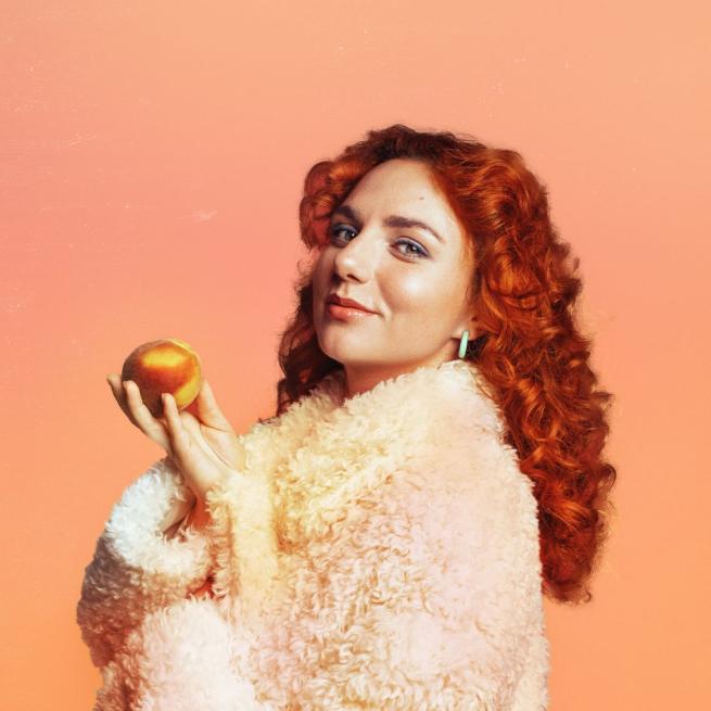 Ania Magliano stands in profile, wearing a fluffy coat and holding a peach in her hand. She smiles against a plain peach background.