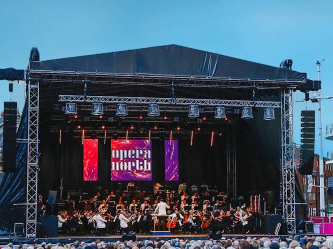 The orchestra of Opera North performing in Millenium Square on stage in front of a large crowd, under a banner with the words 'Opera North'.