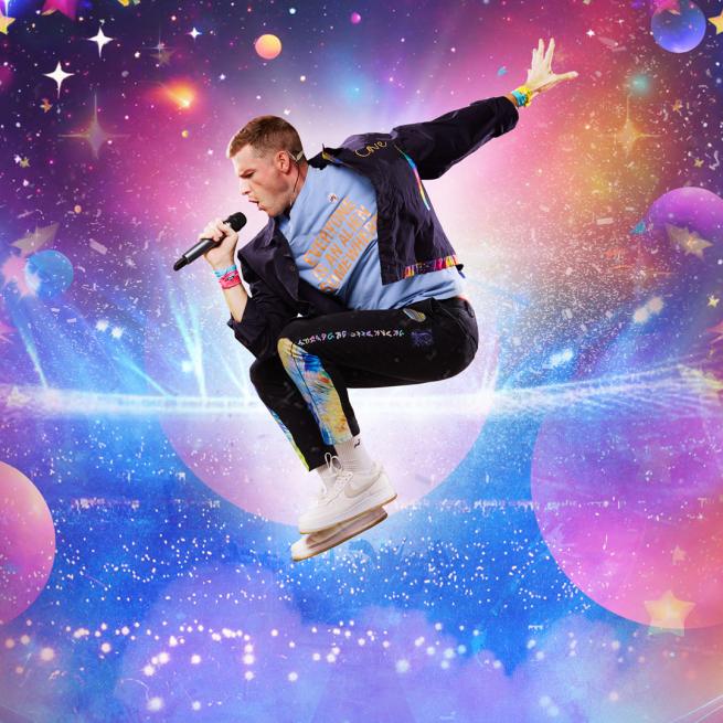 A man in a jacket, blue t-shirt and colourful wristbands jumps into the air and sings into a microphone, surrounded by a colourful galaxy of blue, pink and purple planets.