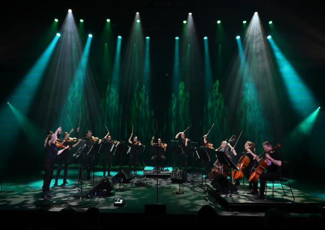 The image shows a group of musicians on a dimly lit stage, arranged in a semicircle and playing string instruments under dramatic green and white spotlights. Cables, music stands, and microphones surround the performers, creating the setup for a live orchestral performance.
