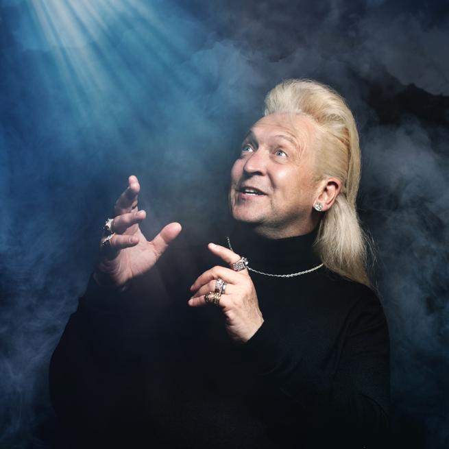 Clinton Baptiste in a black turtle neck gesturing to the sky surrounded by stage smoke.