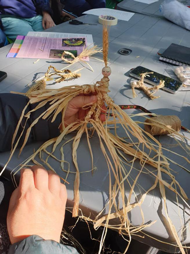 image shows a hand holding a corn doll, made with plated rafia, with a round wooden face and a dried ear of corn headdress.