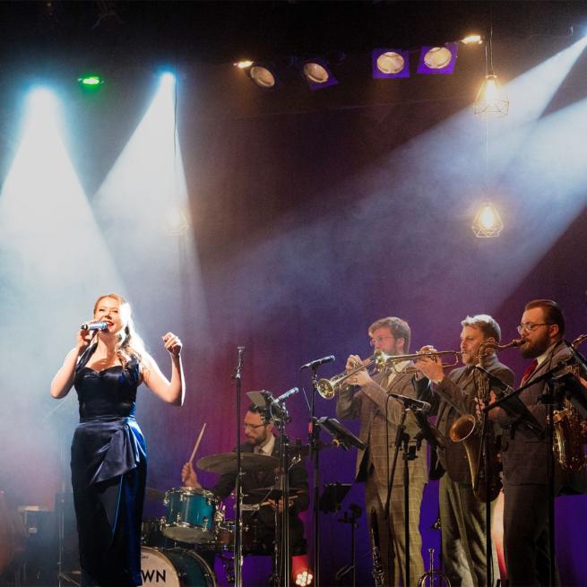 From left to right, a stage is filled with suited musicians: a drummer, two trumpet players and a saxophonist. In the foreground is a glamorous female vocalist below a hazy spotlight.
