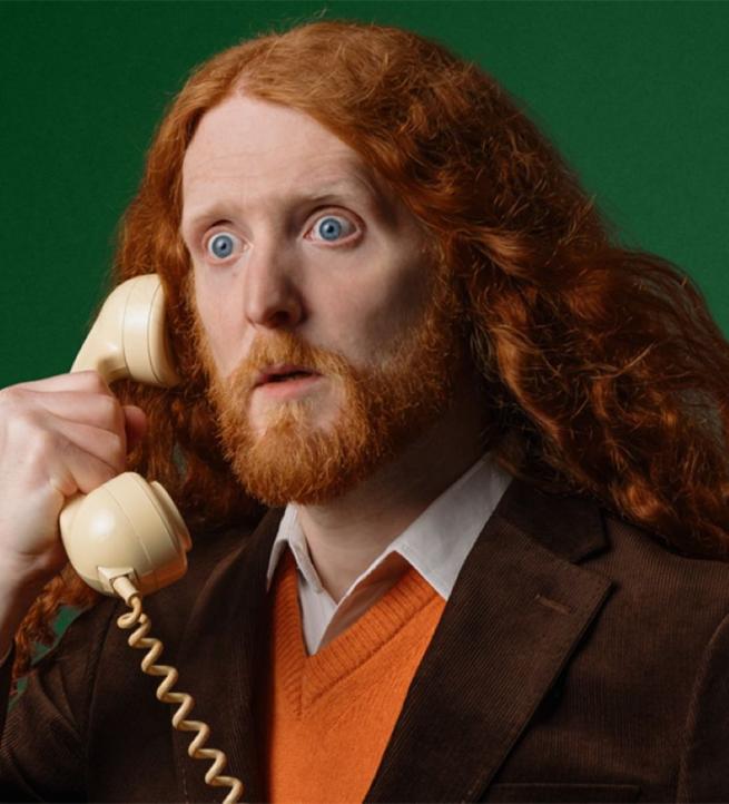 A ginger haired man with hair somewhat redolent of King Charles the Second holds an old fashioned telephone receiver to his ear, his eyes are wide with surprise or shock, it's hard to tell which , he is wearing an brown jacket, orange v neck jumper and a stiff collared white shirt open at the neck