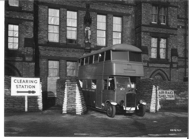 A Second World War scene showing the front of a building with a Routemaster-style bus parked outside. The bus is marked “Mobile First Aid Post” and is surrounded by stacked sandbags. Nearby signs read “Clearing Station” and “Air Raid Shelter,” indicating its use as an emergency medical point during the war.