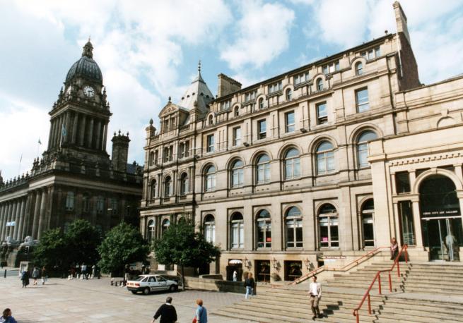 Photograph showing exterior of Leeds Central Library with a paved area in front of it. The clock tower of Leeds Town Hall can be seen behind the library.