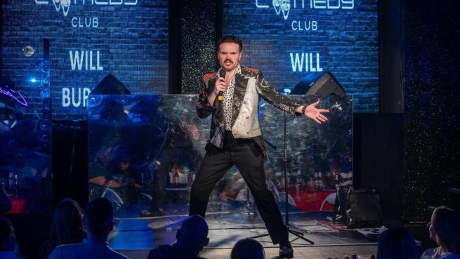 A white man with dark hair and a moustache poses with a microphone against a blue brick background.