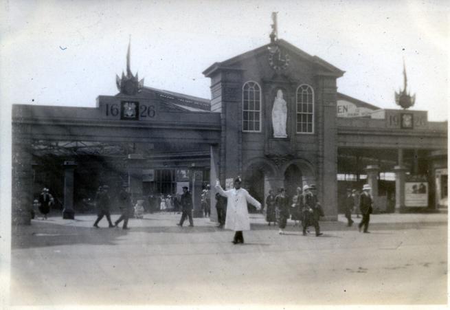 An image of Leeds City Square in 1926, with model moot hall erected for the tercentenary celebrations of Leeds getting its charter. A large building facade with a white statue in a central alcove (Queen Anne) stands in the centre. On either side the dates 1626 and 1926 are etched in what looks like stone. This is entrance to a station concourse. Pedestrians are milling about in period 1920s dress. A policeman in a white coat and gloves stands beneath the arch directing people.