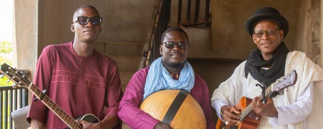 Three men standing together, holding guitars and a traditional instrument outdoors.