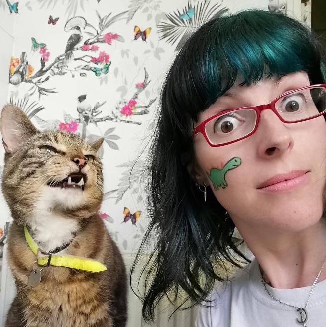Nicky Bray, a woman with dark hair wearing red glasses, with face paint on, next to a cat