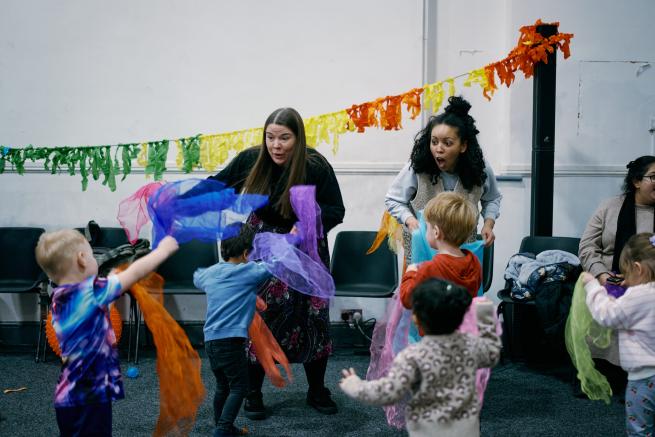 Children play energetically with colorful scarves in a lively room. Two women join, expressing joy and encouragement. Bright decorations adorn the space.