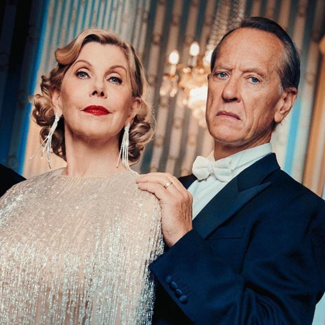 With serious expressions, Christine Baranski wears dangly earrings and a shimmering dress, standing beside Richard E Grant in a dark tuxedo with a white bowtie. A chandelier is visible by a window and patterned wallpaper behind them.