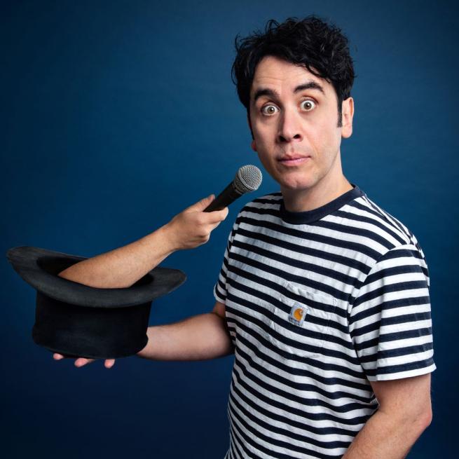 In a striped t-shirt, Pete Firman displays a shocked expression to the camera as an arm holding a microphone emerges from a top hat he is holding.