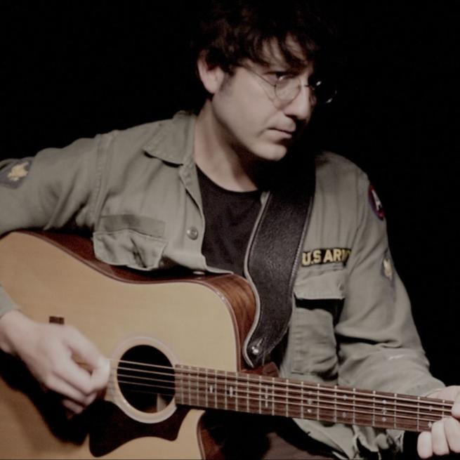 A John Lennon tribute artist strums an acoustic guitar, wearing glasses and a khaki US Army bomber jacket.