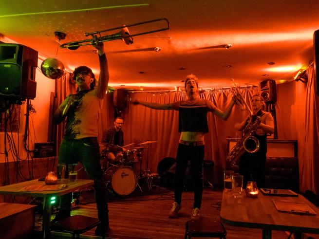 A band with trumpets, trombones and drums plays on the red lit stage.