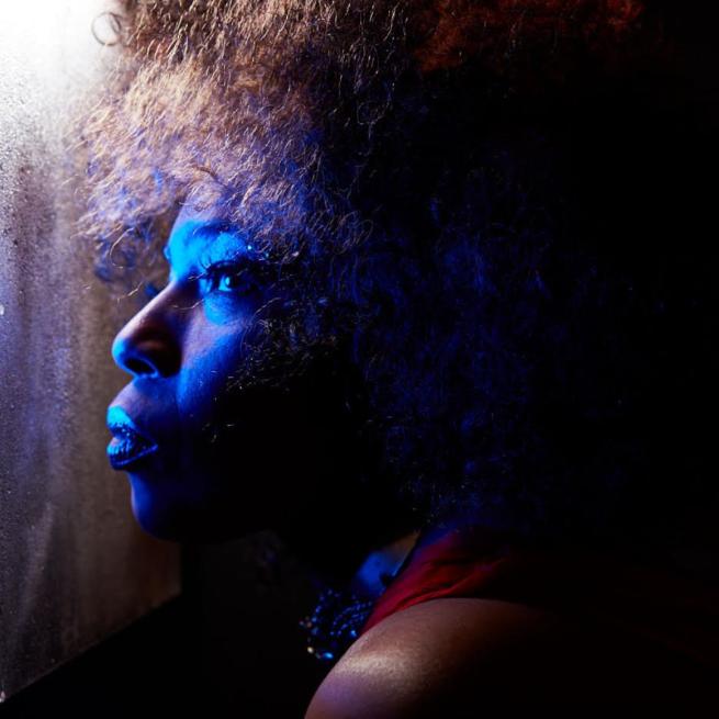 Shot in profile, Macy Gray looks through a misty window, her face lit by a deep blue light, the rest of her in shadows.
