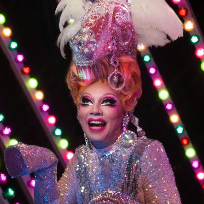 Ginger Johnson smiles in drag makeup and a glittery, bedazzled pony outfit in front of colourful LED lights.