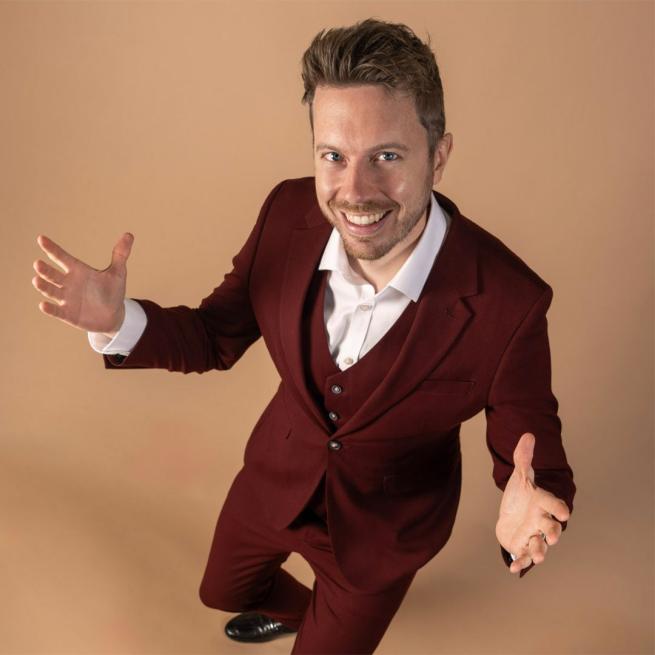 James B Partridge smiles in a maroon suit, with arms open.