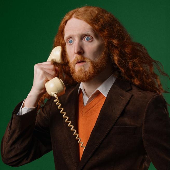 Alasdair Beckett-King's long red hair breezes over his shoulder as he holds a white telephone to his ear with a shocked expression. He wears an orange jumper and brown suit jacket, as he stands in front of a dark green background.