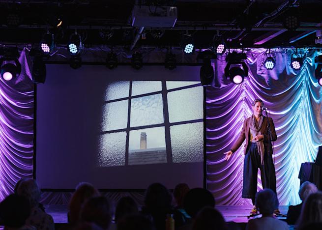 Artist Hannah Lamb standing on the stage of the Old Woollen, with purple and blue lights glowing against a ruched backdrop. She is standing in front of a microphone stand whilst gesturing toward a projector showing an image of a Mill chimney through an old window.