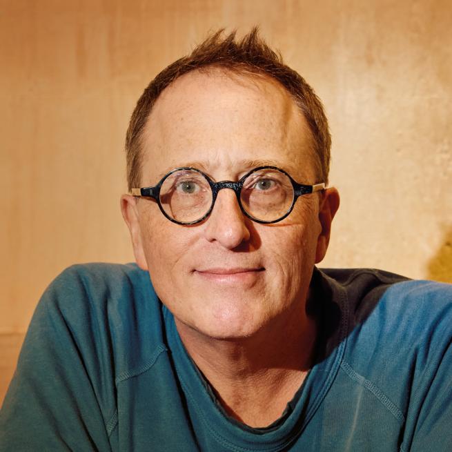 Jon Ronson smiles in a relaxed pose against a bare wall.